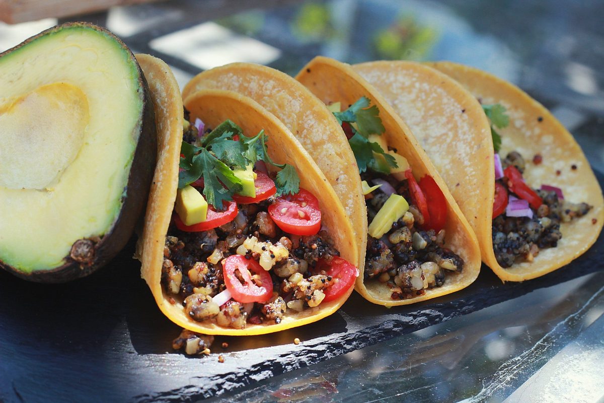 Three corn tortilla tacos filled with browned tempeh crumbles, tomato, chili and cilantro, on a slate board beside a halved avocado