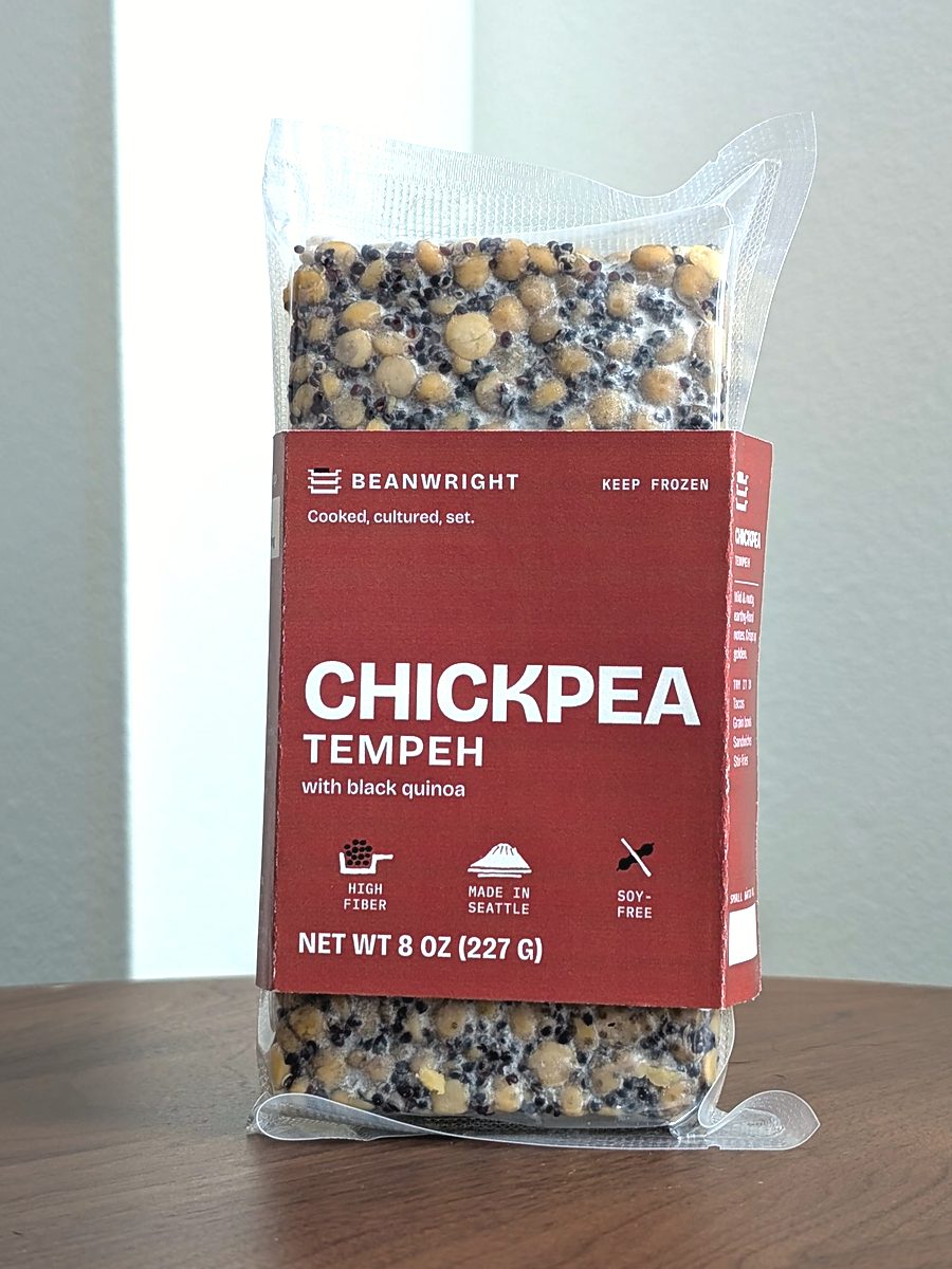 Chickpea tempeh in its burnt-ochre band. Pale chickpeas and black quinoa show through the vacuum pack above and below.