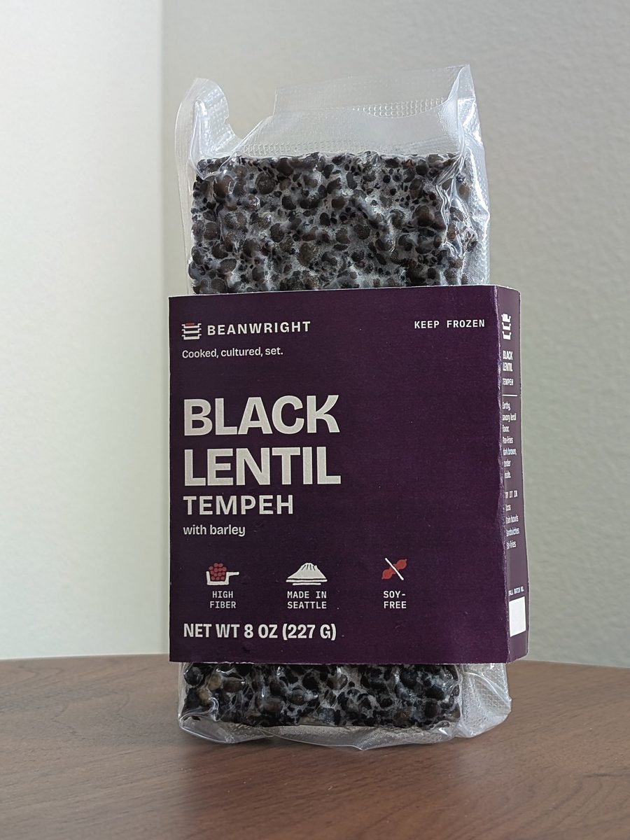 Black lentil tempeh in its aubergine band. Black lentils bound in white culture show through the vacuum pack above and below.