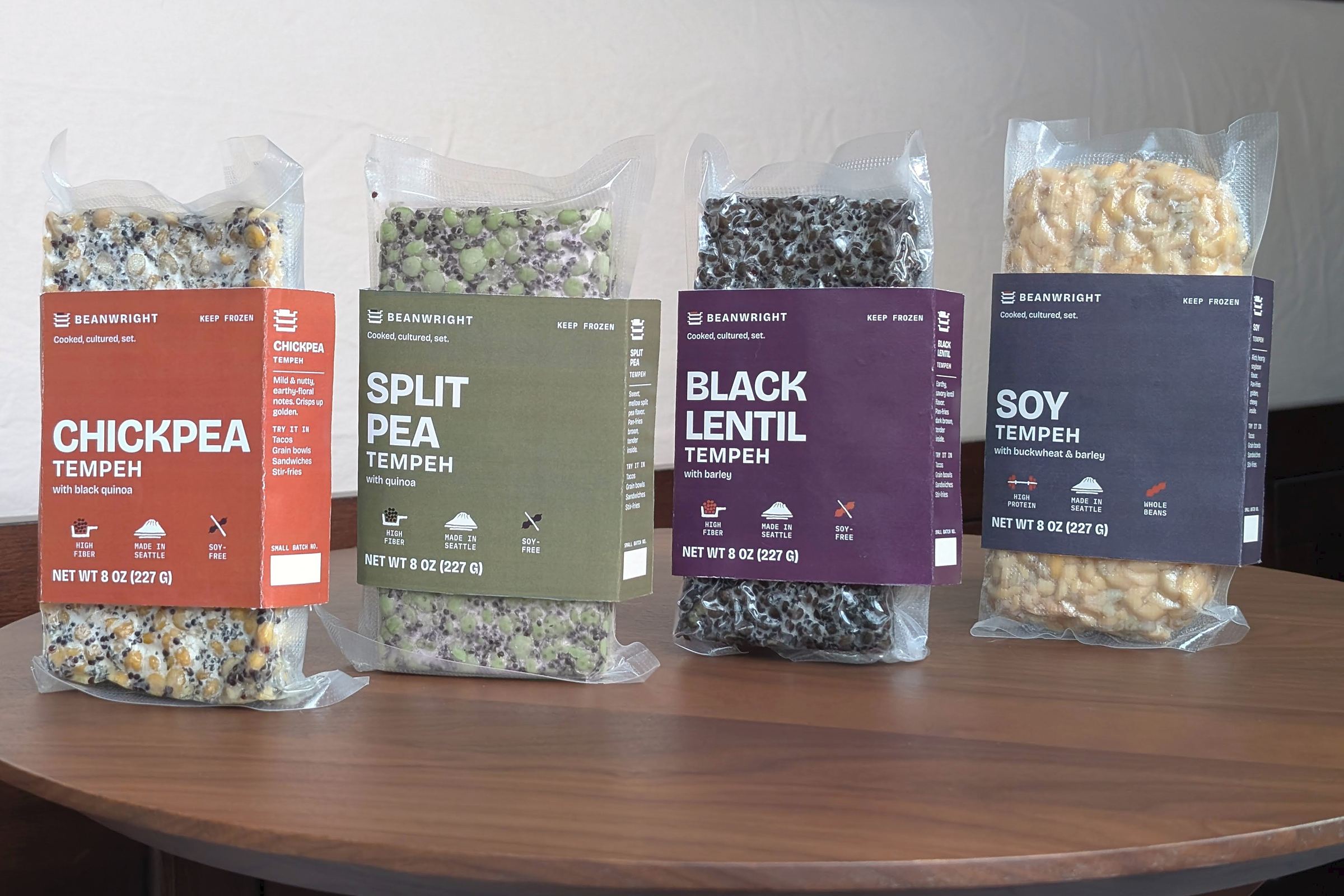 Four Beanwright tempeh packs standing in a row on a wooden table: chickpea in a burnt-ochre paper band, split pea in olive, black lentil in aubergine and soy in slate. Each block shows through its vacuum pack above and below the band.